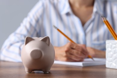 Photo of Woman taking notes at wooden table, focus on piggy bank