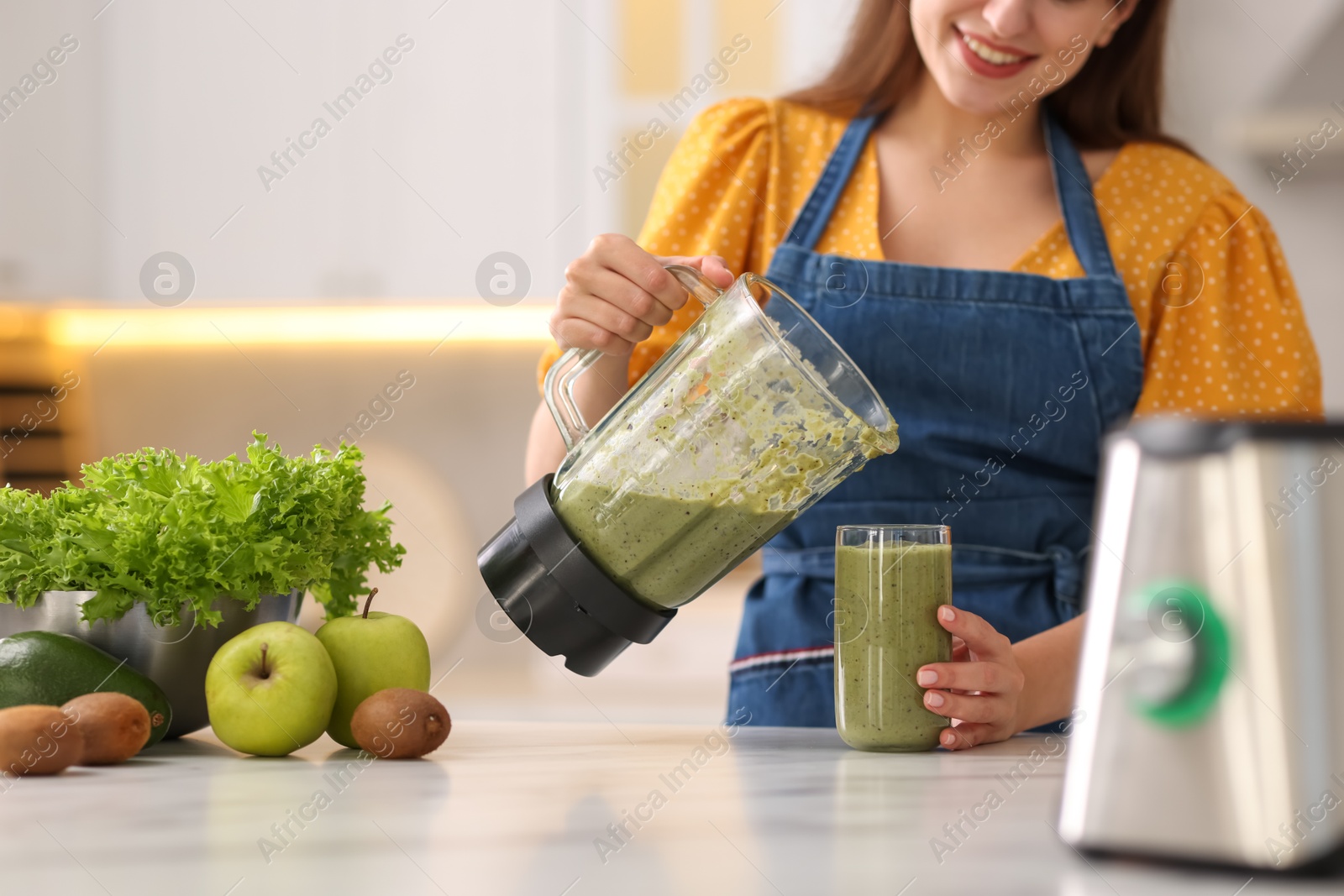 Photo of Woman pouring fresh smoothie from blender cup into glass at white marble table in kitchen, closeup