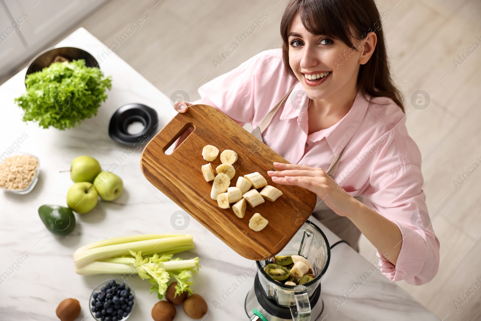 Photo of Young woman making delicious smoothie with blender at white marble table in kitchen, above view