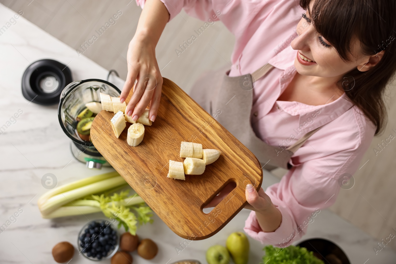 Photo of Young woman making delicious smoothie with blender at white marble table in kitchen, top view