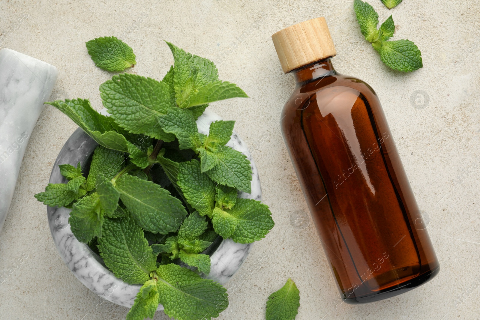 Photo of Bottle of essential oil, mint, mortar and pestle on light textured table, flat lay