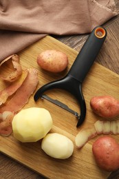 Fresh raw potatoes, peels and peeler on wooden table, flat lay