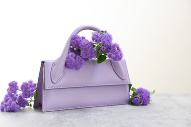 Photo of Stylish violet bag and ageratum flowers on gray table against light background, space for text