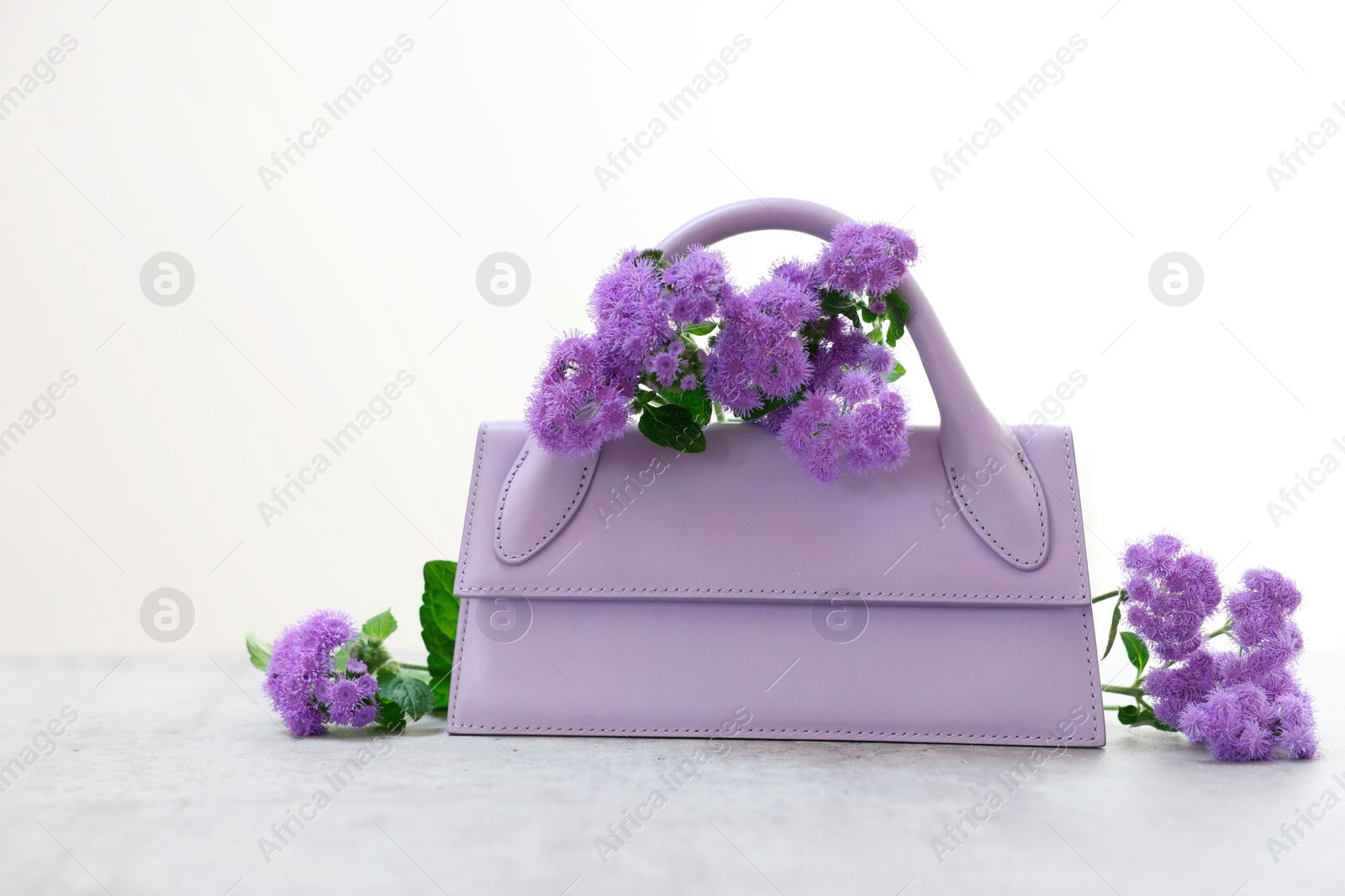 Photo of Stylish violet bag and ageratum flowers on gray table against light background, space for text