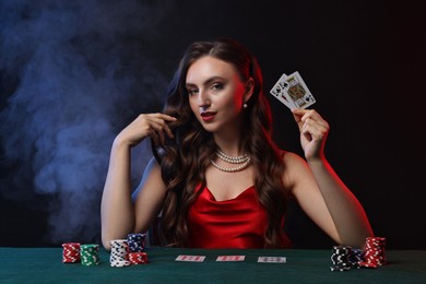 Charming woman playing poker at table on black background with smoke