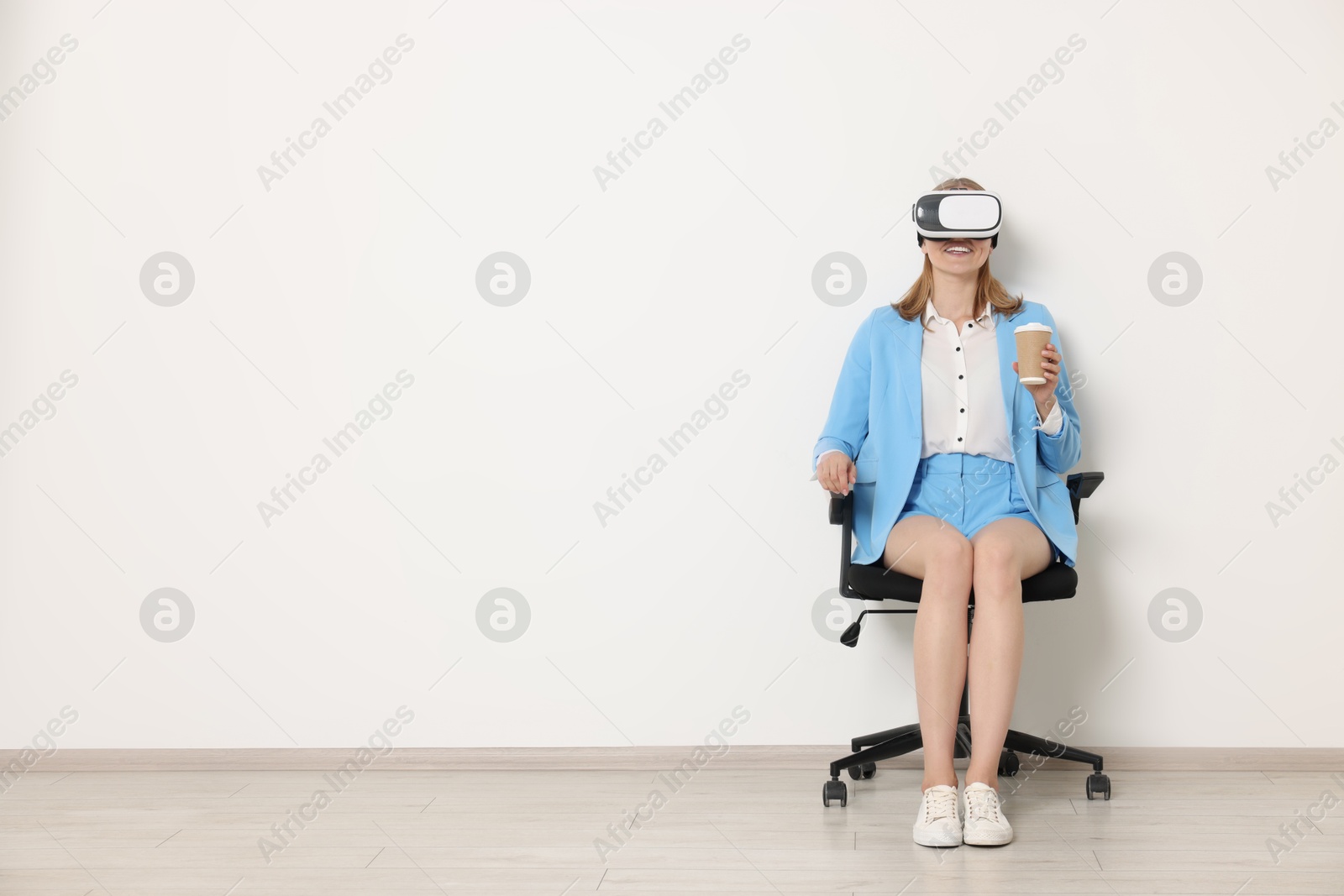 Photo of Happy woman with virtual reality headset and paper cup sitting on chair indoors, space for text