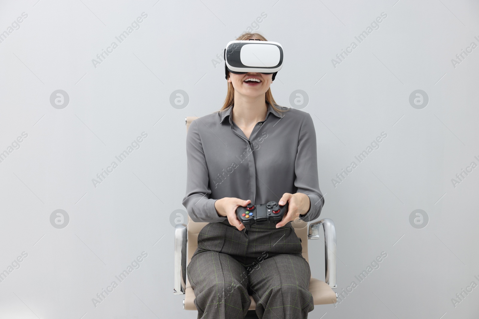 Photo of Happy woman with virtual reality headset and controller sitting on chair against gray background