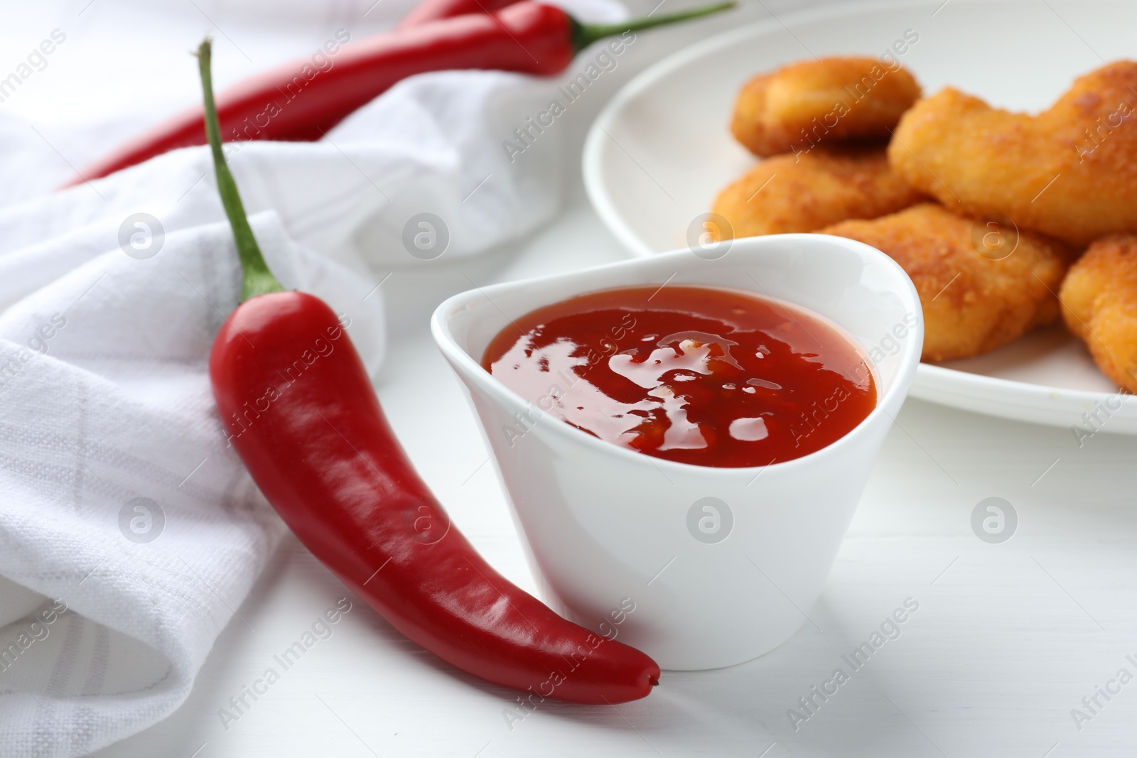 Photo of Spicy chili sauce in bowl and fresh peppers on white wooden table, closeup