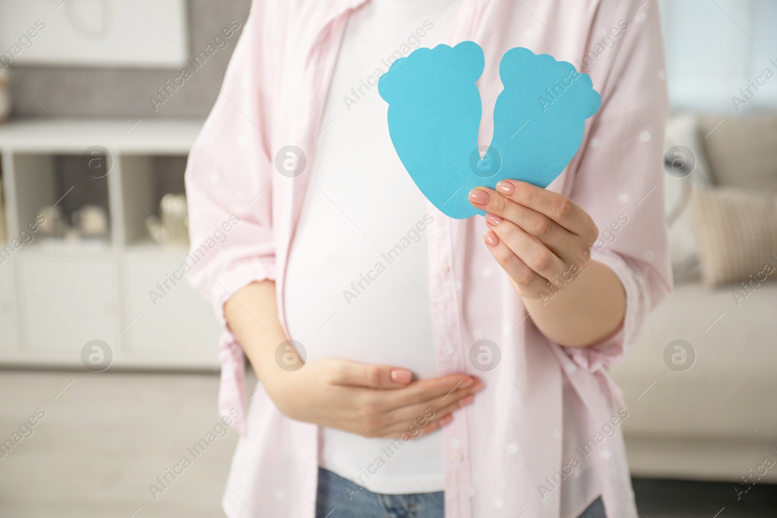 Photo of Expecting twins. Pregnant woman holding two paper cutouts of feet at home, closeup