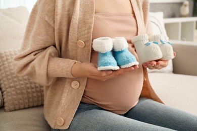 Photo of Expecting twins. Pregnant woman holding two pairs of shoes at home, closeup