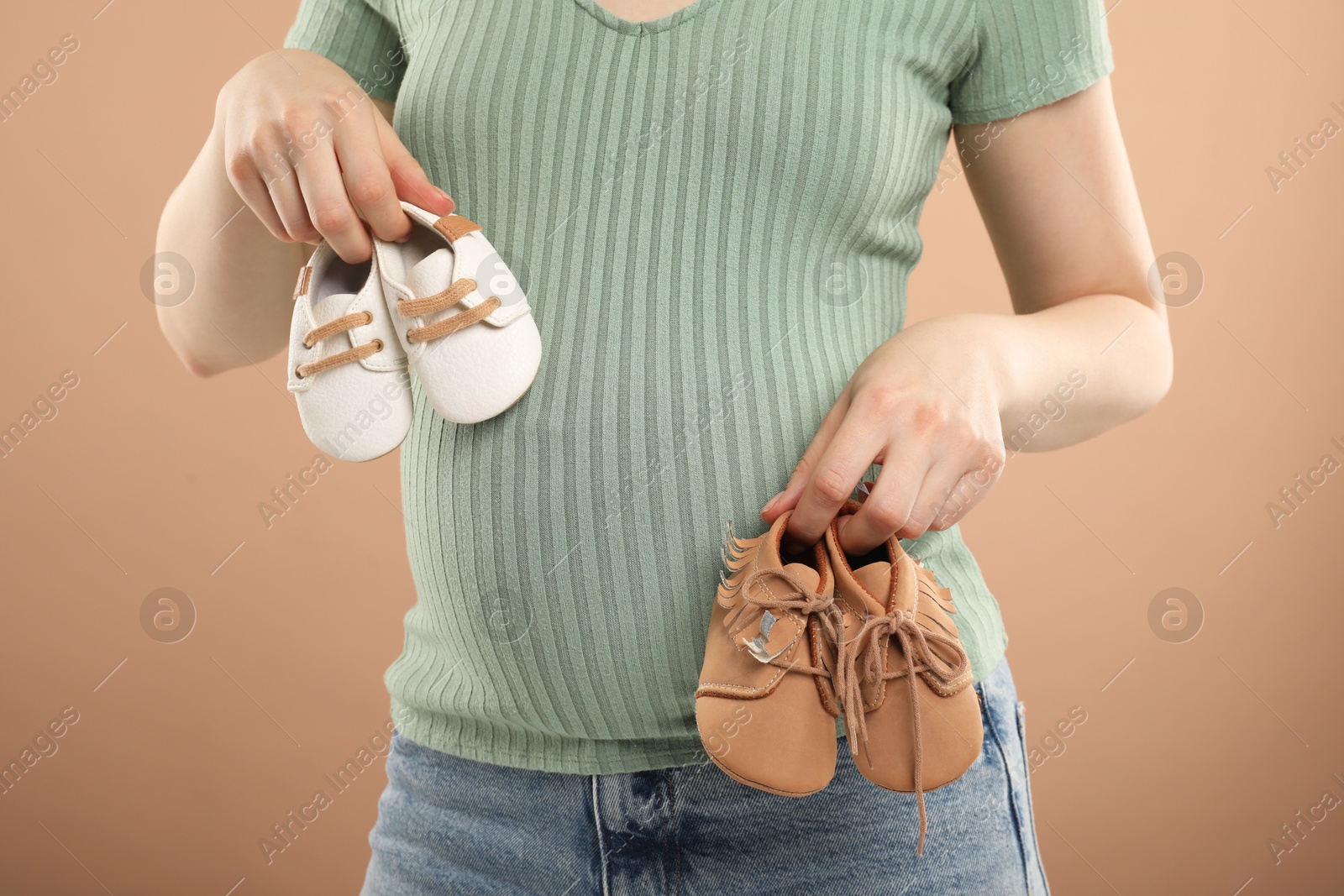 Photo of Expecting twins. Pregnant woman holding two pairs of shoes on light brown background, closeup