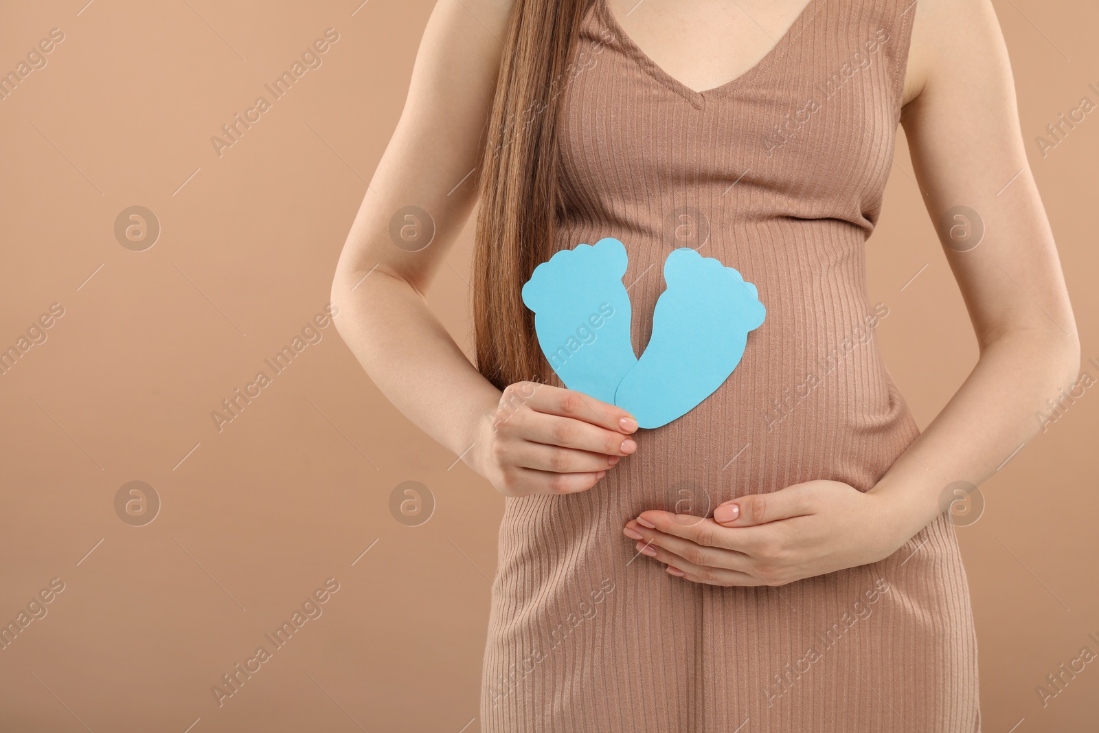 Photo of Expecting twins. Pregnant woman holding two paper cutouts of feet on light brown background, closeup. Space for text