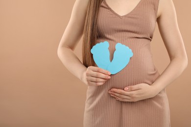 Expecting twins. Pregnant woman holding two paper cutouts of feet on light brown background, closeup. Space for text