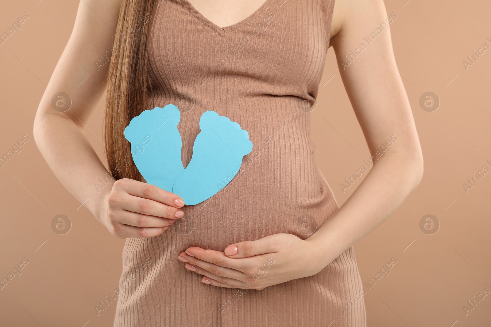 Photo of Expecting twins. Pregnant woman holding two paper cutouts of feet on light brown background, closeup