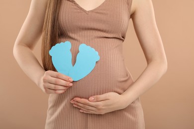 Photo of Expecting twins. Pregnant woman holding two paper cutouts of feet on light brown background, closeup