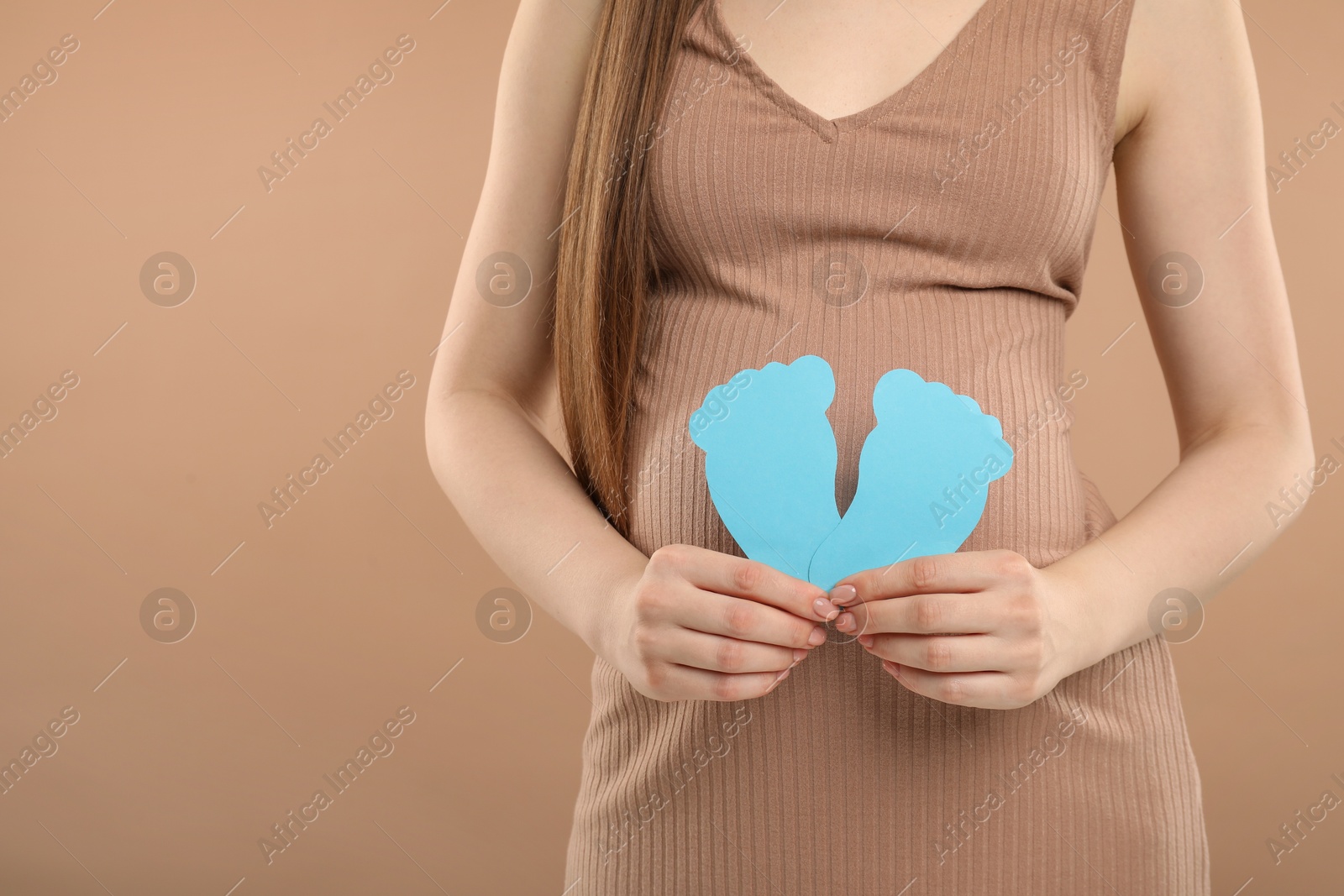 Photo of Expecting twins. Pregnant woman holding two paper cutouts of feet on light brown background, closeup. Space for text