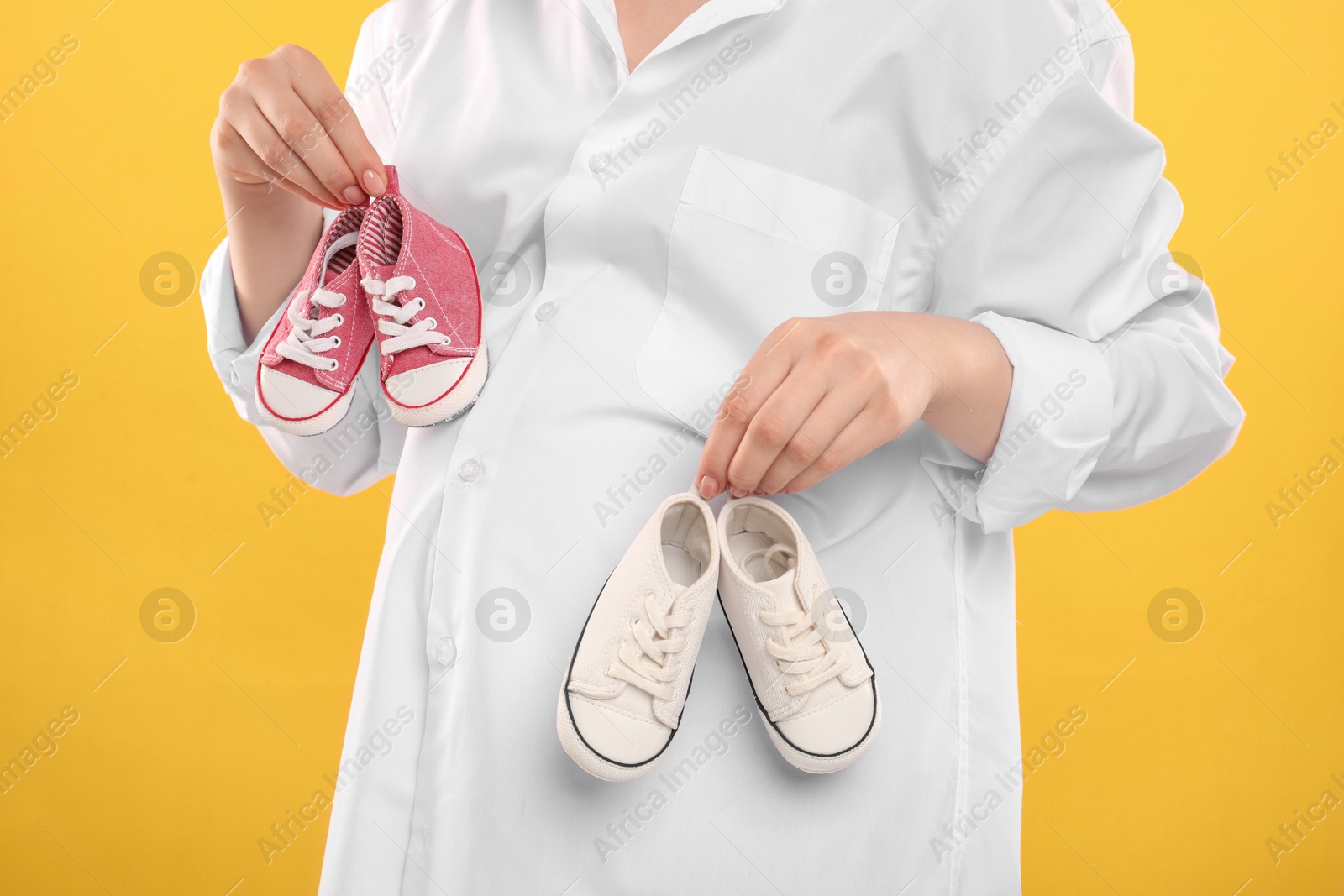 Photo of Expecting twins. Pregnant woman holding two pairs of shoes on yellow background, closeup