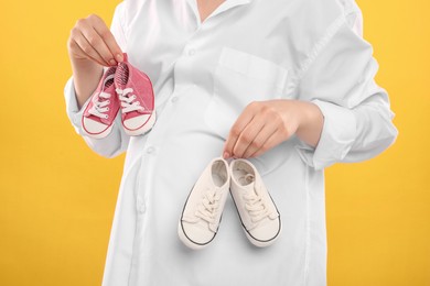 Expecting twins. Pregnant woman holding two pairs of shoes on yellow background, closeup