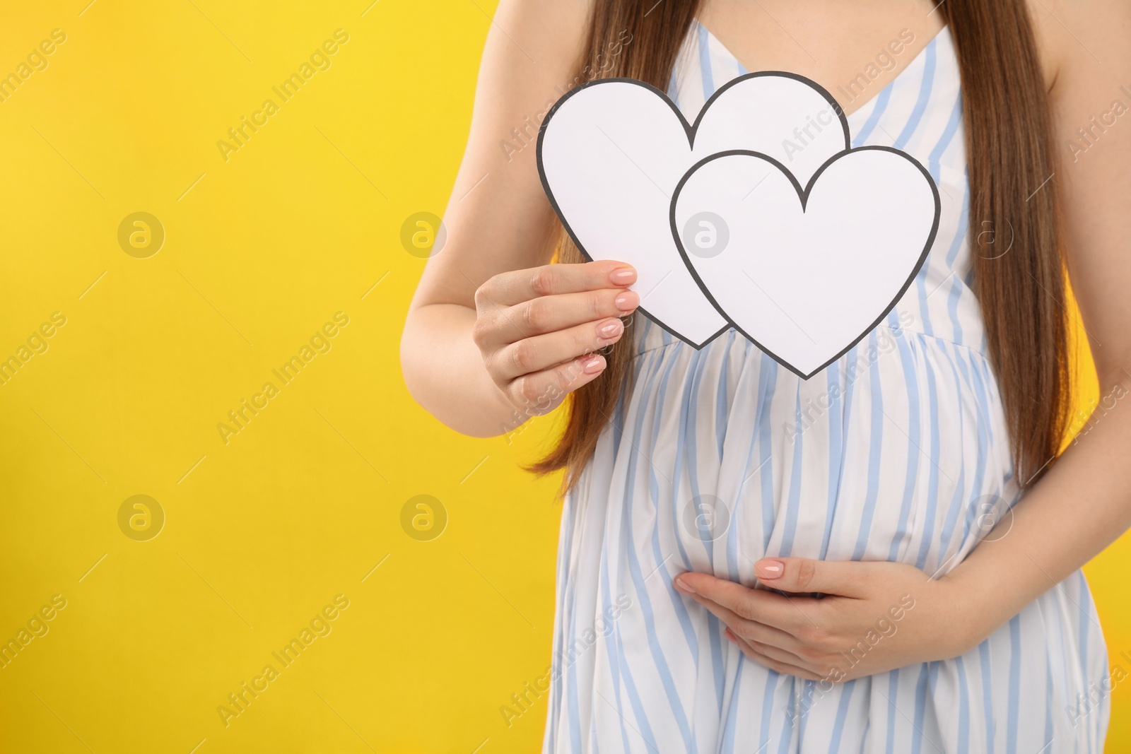 Photo of Expecting twins. Pregnant woman holding two paper cutouts of hearts on yellow background, closeup. Space for text