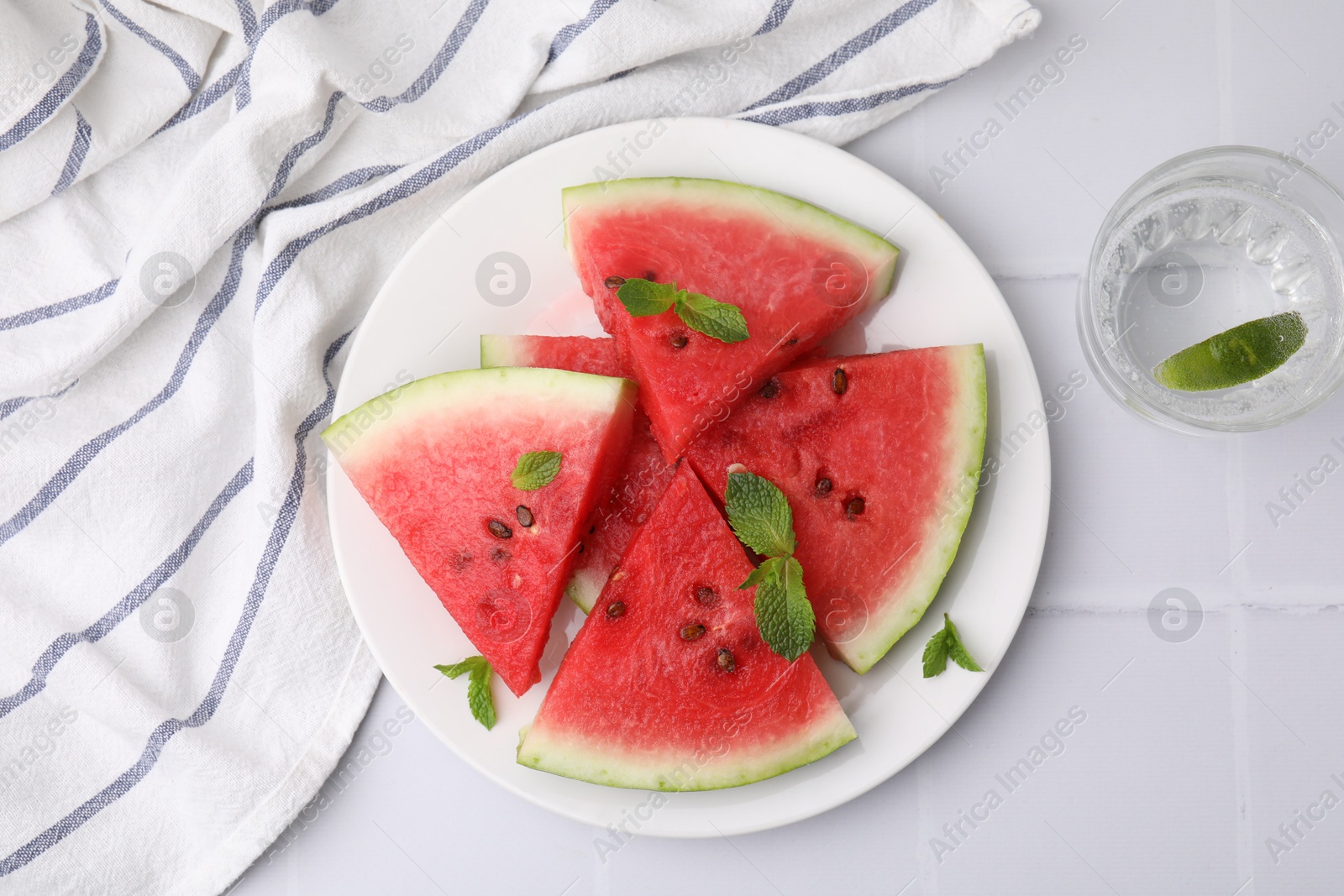 Photo of Fresh watermelon slices with mint leaves and glass of water on white tiled table, flat lay