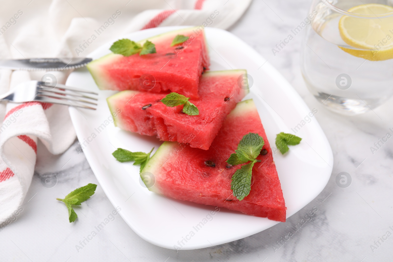 Photo of Fresh watermelon slices with mint leaves and glass of water on white marble table, closeup