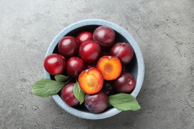 Photo of Fresh plums and leaves in bowl on grey textured table, top view