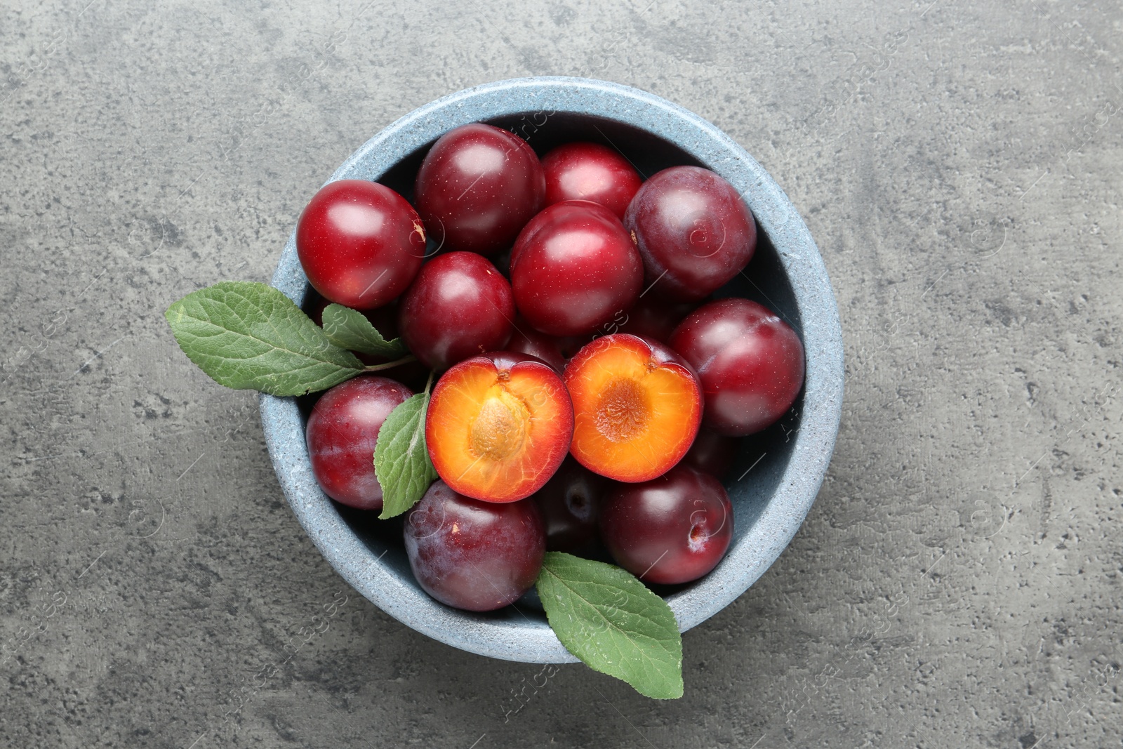 Photo of Fresh plums and leaves in bowl on grey textured table, top view