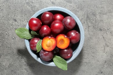 Photo of Fresh plums and leaves in bowl on grey textured table, top view