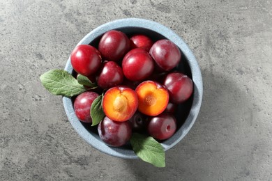 Photo of Fresh plums and leaves in bowl on grey textured table, top view