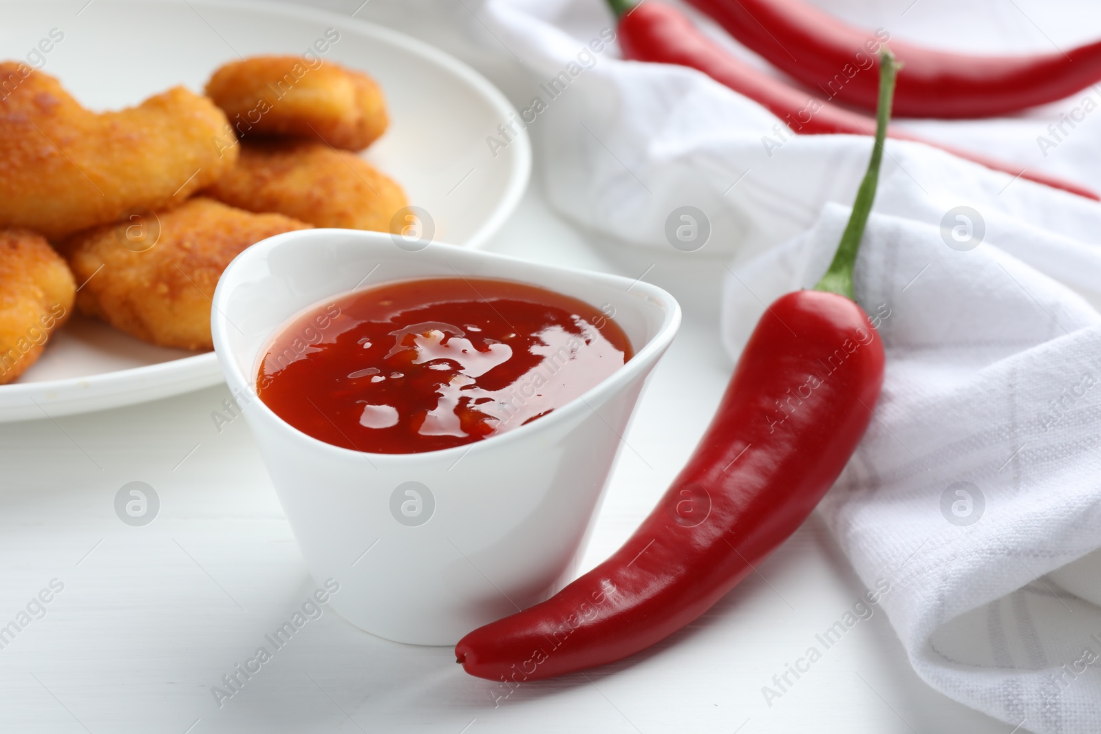 Photo of Spicy chili sauce in bowl and fresh peppers on white wooden table, closeup