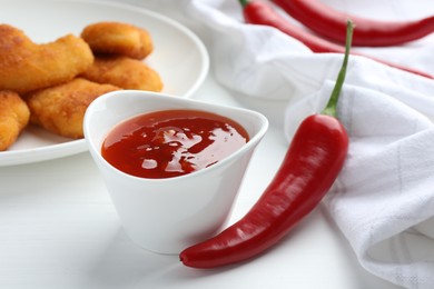 Photo of Spicy chili sauce in bowl and fresh peppers on white wooden table, closeup