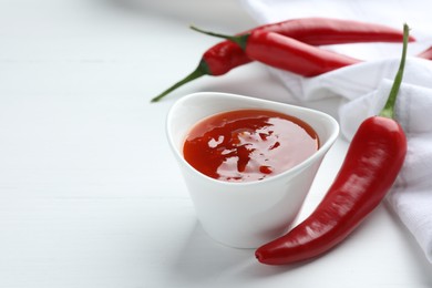 Photo of Spicy chili sauce in bowl and fresh peppers on white wooden table, space for text