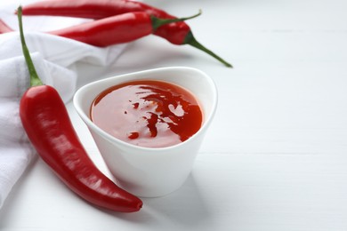 Photo of Spicy chili sauce in bowl and fresh peppers on white wooden table, space for text