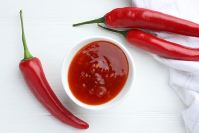 Photo of Spicy chili sauce in bowl and fresh peppers on white wooden table, flat lay