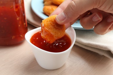 Photo of Woman dipping tasty chicken nugget into chili sauce at wooden table, closeup