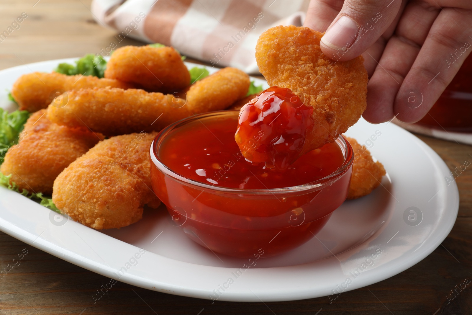 Photo of Woman dipping tasty chicken nugget into chili sauce at wooden table, closeup