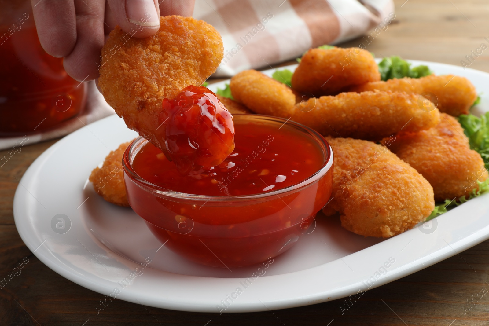 Photo of Woman dipping tasty chicken nugget into chili sauce at wooden table, closeup