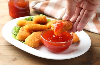 Photo of Woman dipping tasty chicken nugget into chili sauce at wooden table, closeup