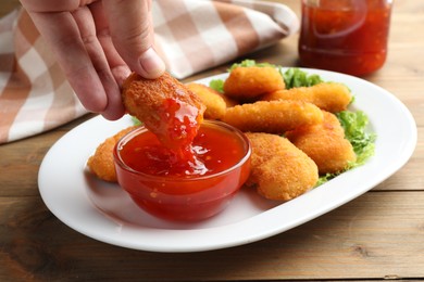 Photo of Woman dipping tasty chicken nugget into chili sauce at wooden table, closeup