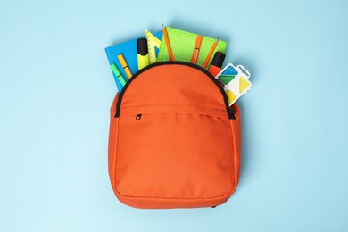 Orange backpack with different school supplies on light blue background, top view