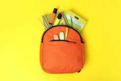 Orange backpack and different school supplies on yellow background, top view