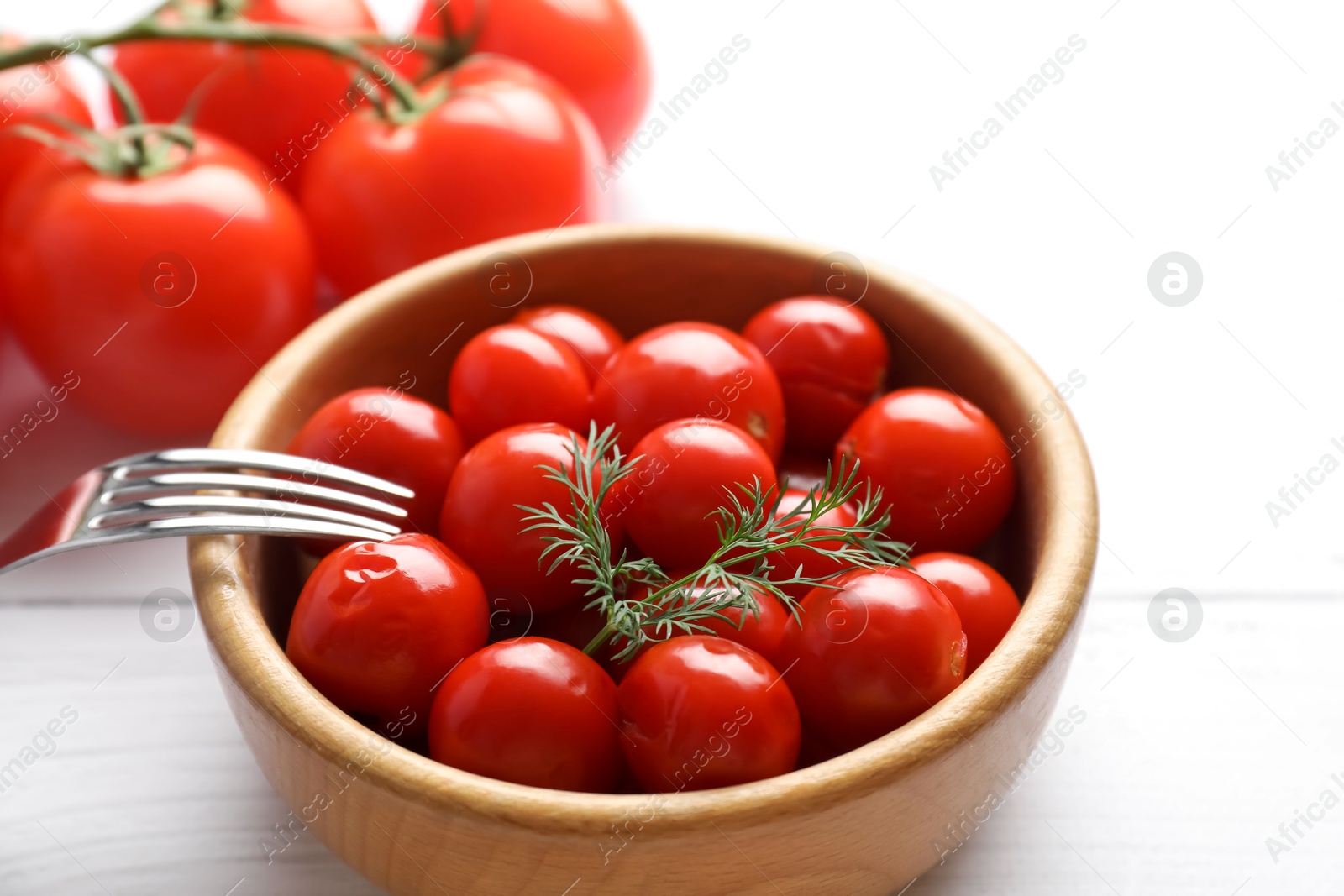 Photo of Tasty pickled tomatoes in bowl, fresh vegetables, dill and fork on white wooden table, closeup