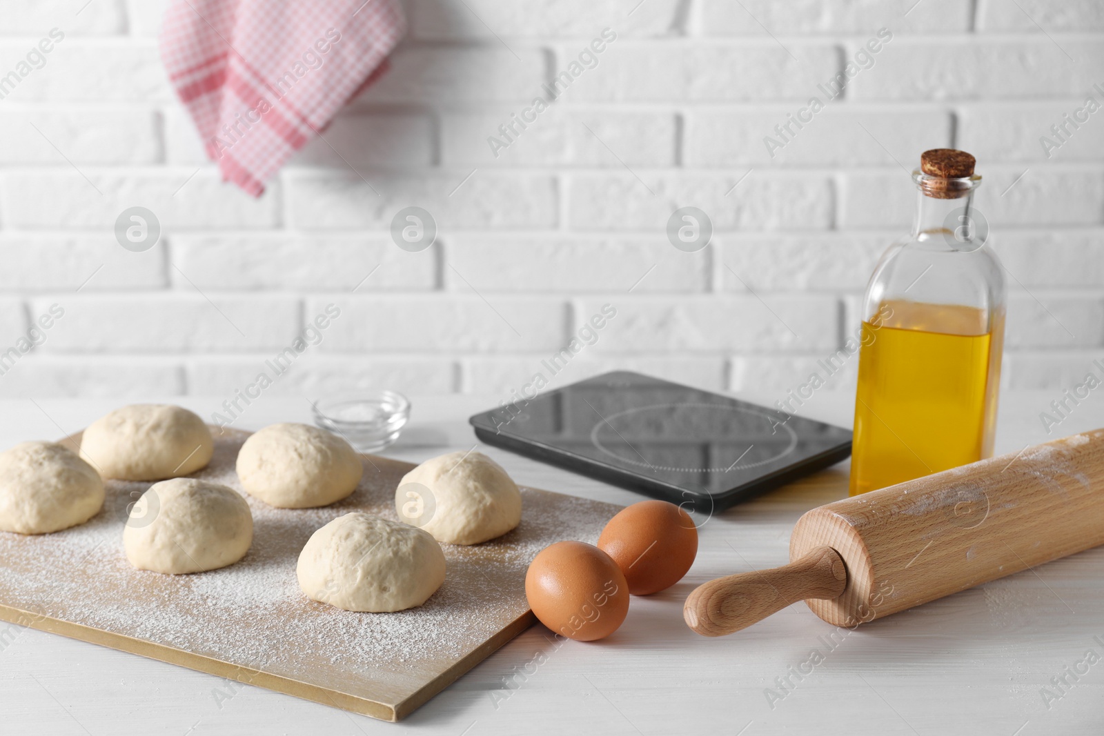 Photo of Raw dough balls, eggs, oil, rolling pin and kitchen scale on white wooden table