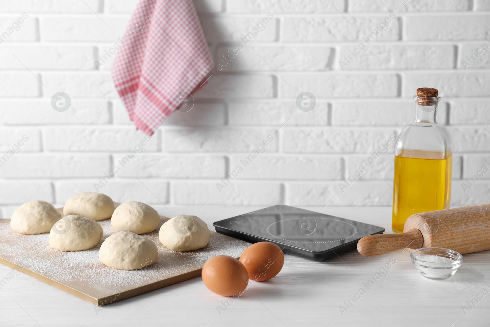 Photo of Raw dough balls, eggs, oil, rolling pin and kitchen scale on white wooden table