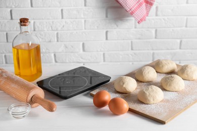 Photo of Raw dough balls, eggs, oil, rolling pin and kitchen scale on white wooden table