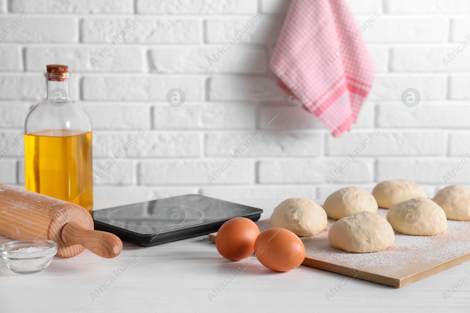 Photo of Raw dough balls, eggs, oil, rolling pin and kitchen scale on white wooden table