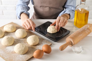 Photo of Woman weighing raw dough ball on kitchen scale at white wooden table, closeup