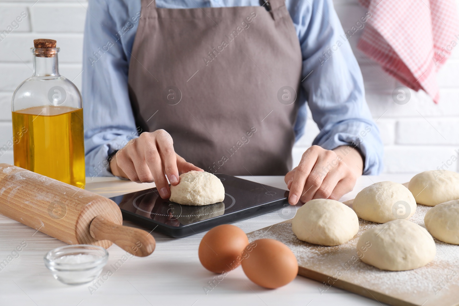 Photo of Woman weighing raw dough ball on kitchen scale at white wooden table, closeup