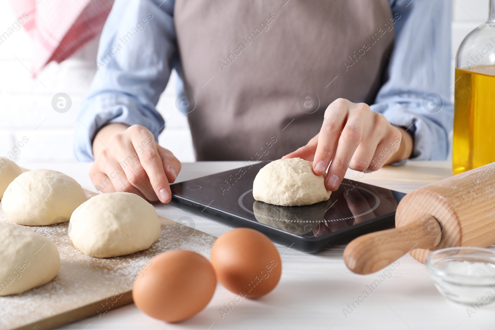 Photo of Woman weighing raw dough ball on kitchen scale at white wooden table, closeup