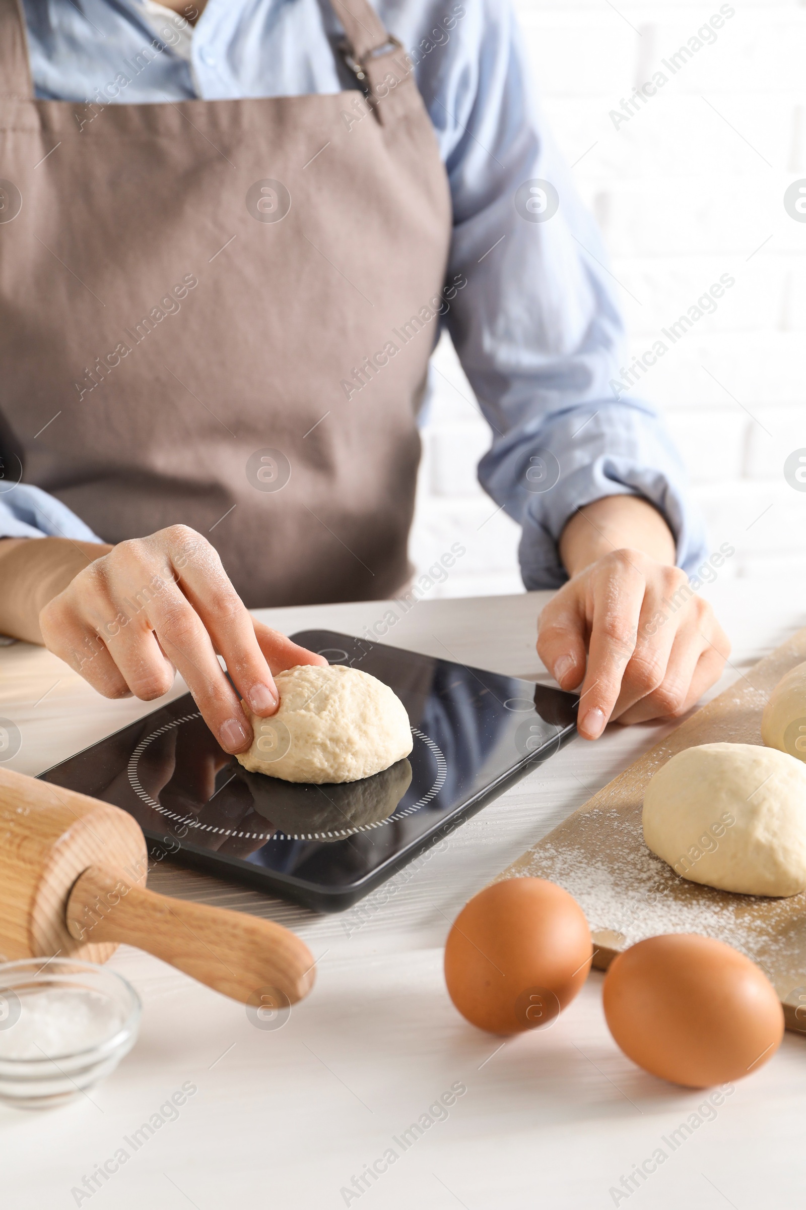 Photo of Woman weighing raw dough ball on kitchen scale at white wooden table, closeup